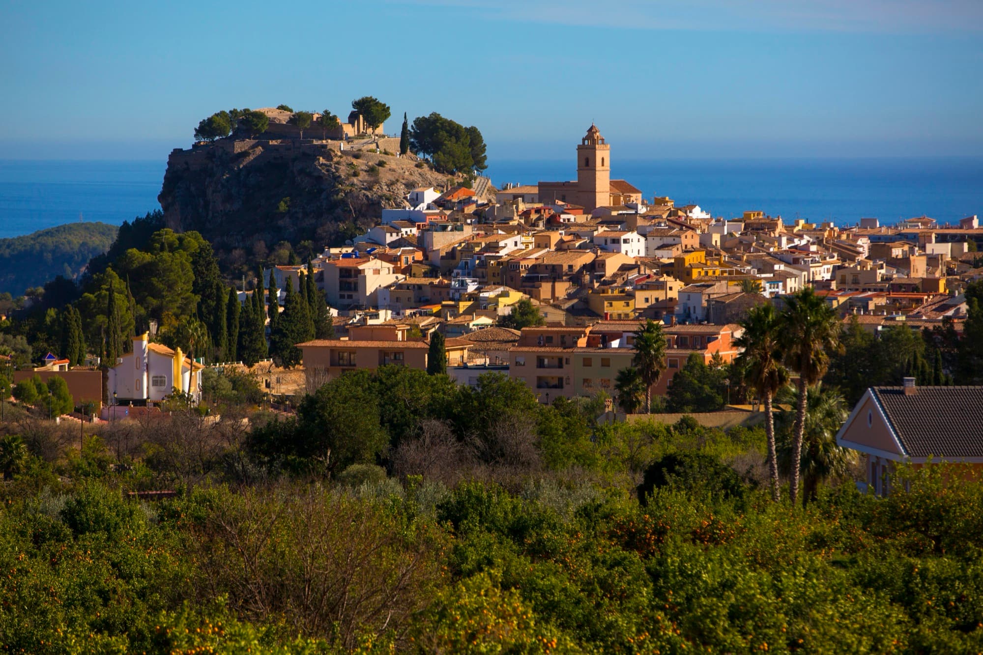 Veduta di Polop con il castello sul colle, il campanile della chiesa e il mare sullo sfondo